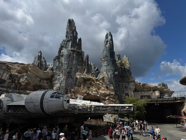 Visitors explore Star Wars: Galaxy’s Edge at Disney’s Hollywood Studios, with the Millennium Falcon in the foreground and Batuu’s rocky spires rising under a blue sky.